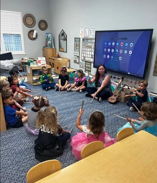 Preschool students sitting in a circle playing rhythm sticks with their teacher.